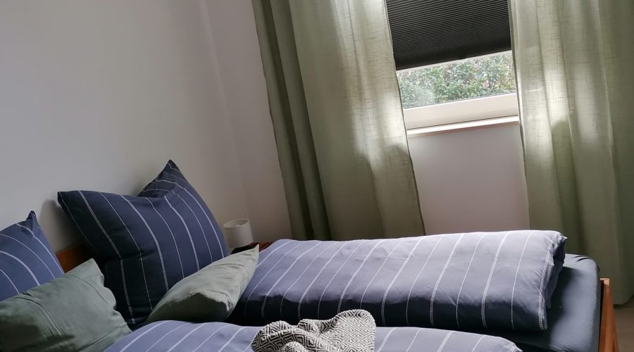 Schlafzimmer mit braunem Doppelbett aus Holz und blauer Bettwäsche mit hellen Streifen. Am Fenster hängen hellgrüne Gardinen.