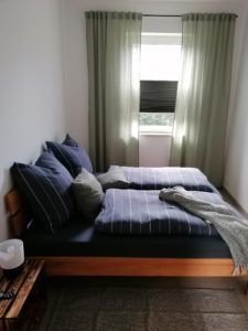 Schlafzimmer in der Ferienwohnung mit braunen Bett und blauer Bettwäsche sowie Fenster mit hellgrünen Vorhängen
