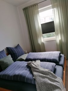 Schlafzimmer in der Ferienwohnung mit braunen Bett und blauer Bettwäsche sowie Fenster mit hellgrünen Vorhängen