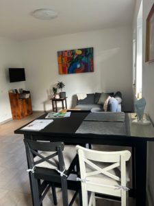 Wohnbereich der Ferienwohnung mit schwarzen Bartisch und Barstühlen, grauen Sofa, Fernseher und farbigen Bild an der Wand